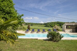 Ein Pool mit sechs Liegestühlen, umgeben von Grünpflanzen und einem Gebäude im Hintergrund.