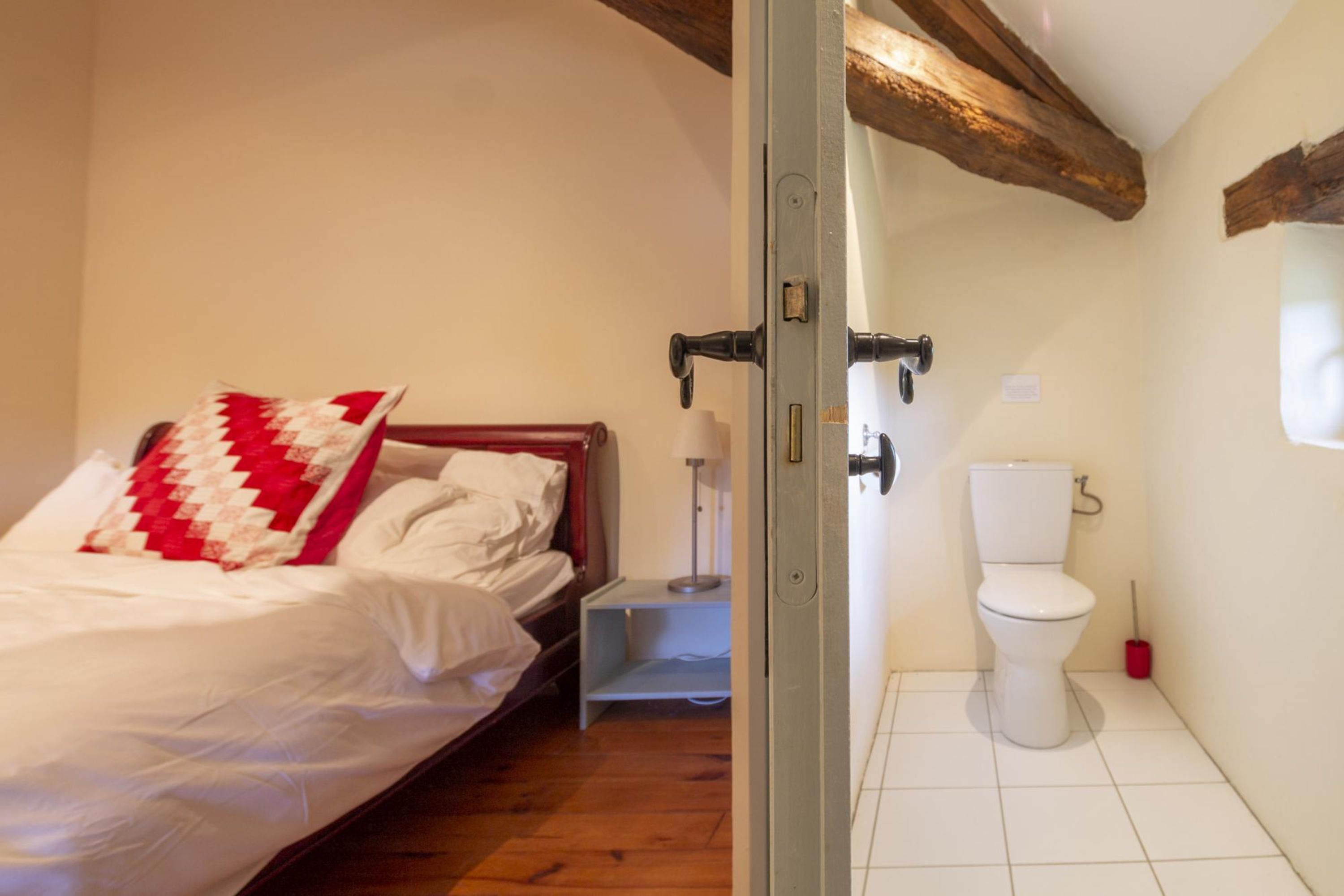 Een slaapkamer met een bed, een nachtkastje, een rood-wit quilt en een wit toilet in de aangrenzende badkamer.