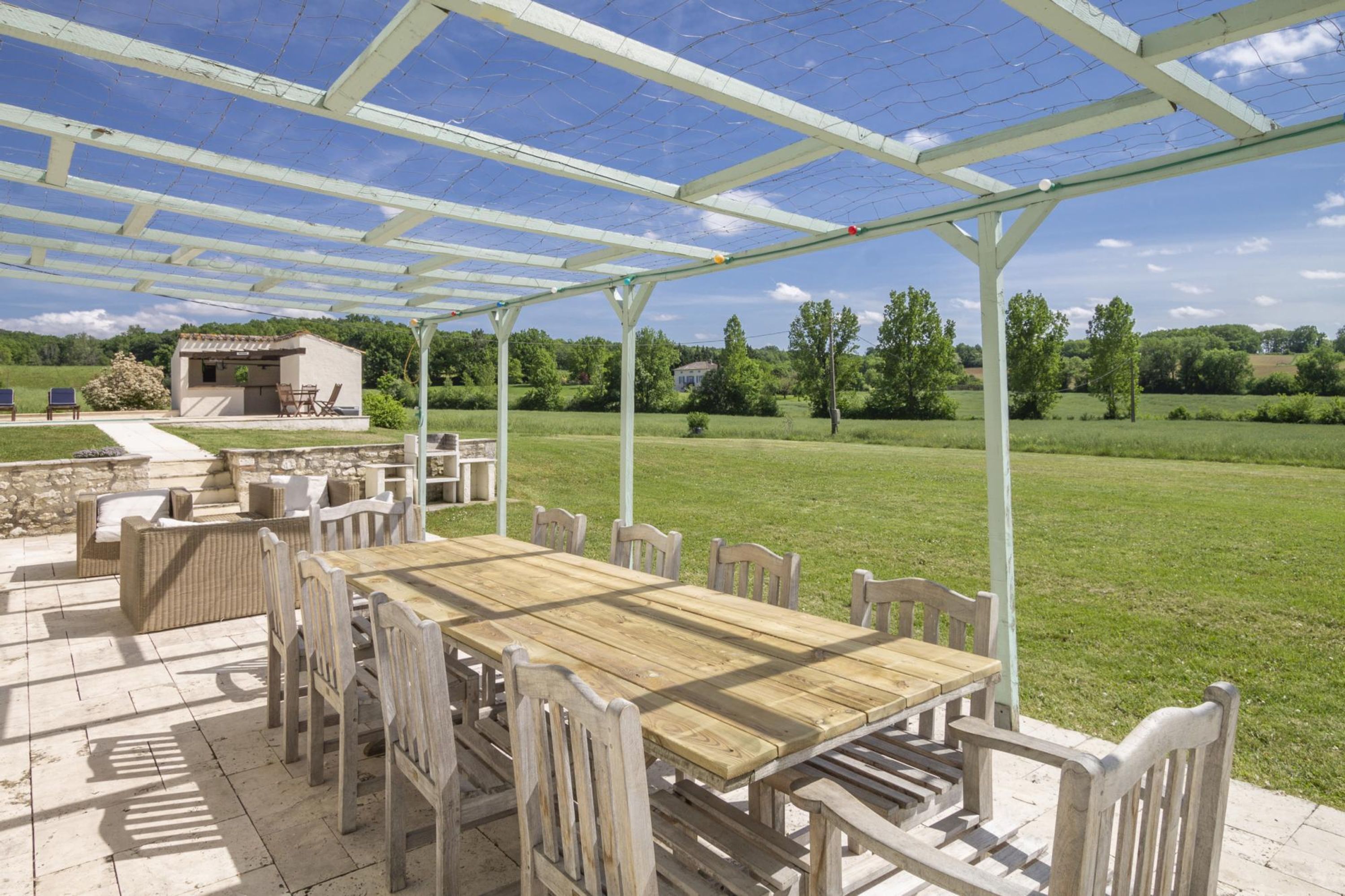 Een terras buiten met een houten tafel en stoelen, een barbecuehoek en een pergola.