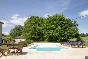 Ein Schwimmbecken mit Liegestühlen und einem Tisch, umgeben von Grünpflanzen und einem blauen Himmel.
