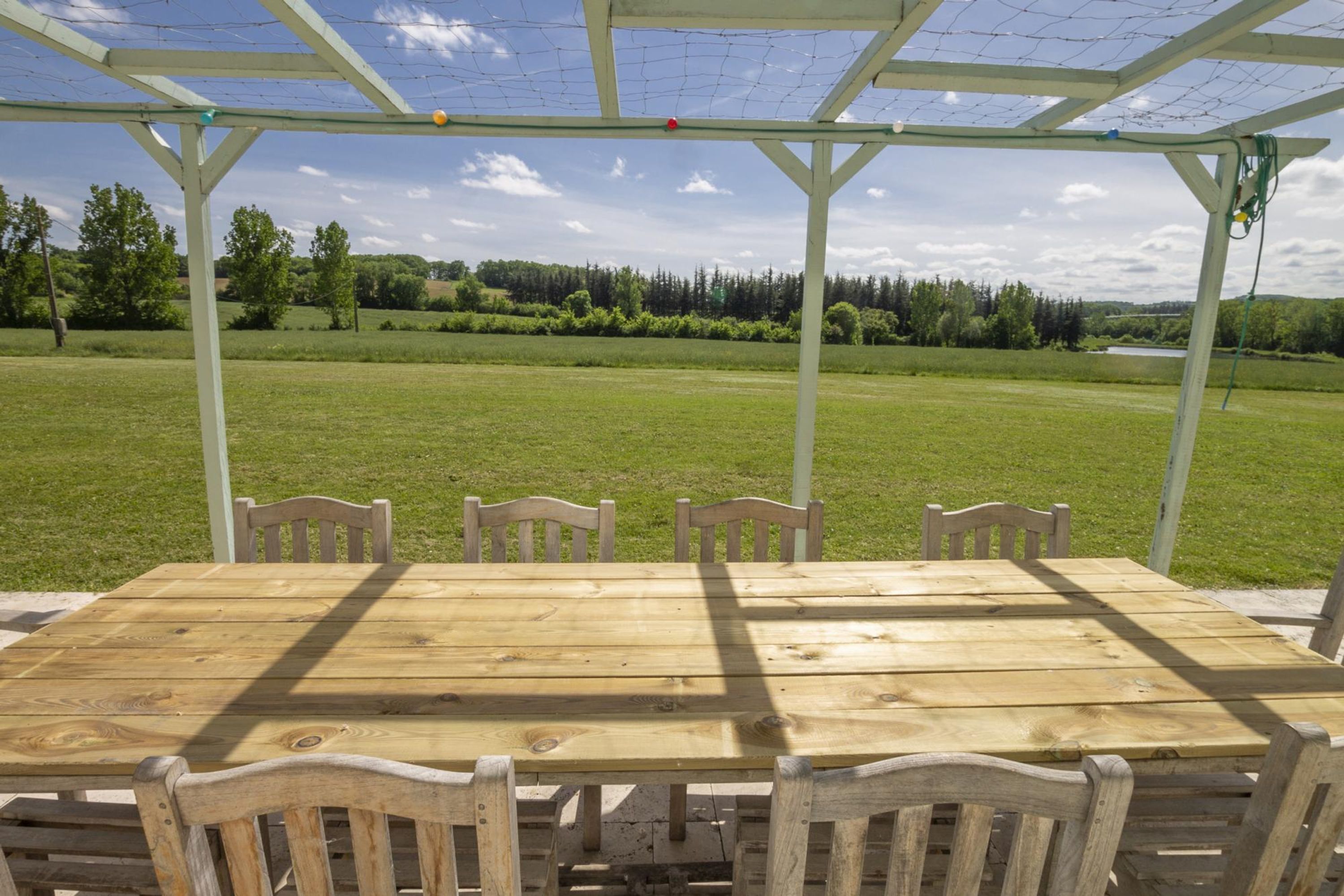 Een houten tafel en stoelen buiten, onder een overdekte ruimte met uitzicht op een groen veld en bomen.
