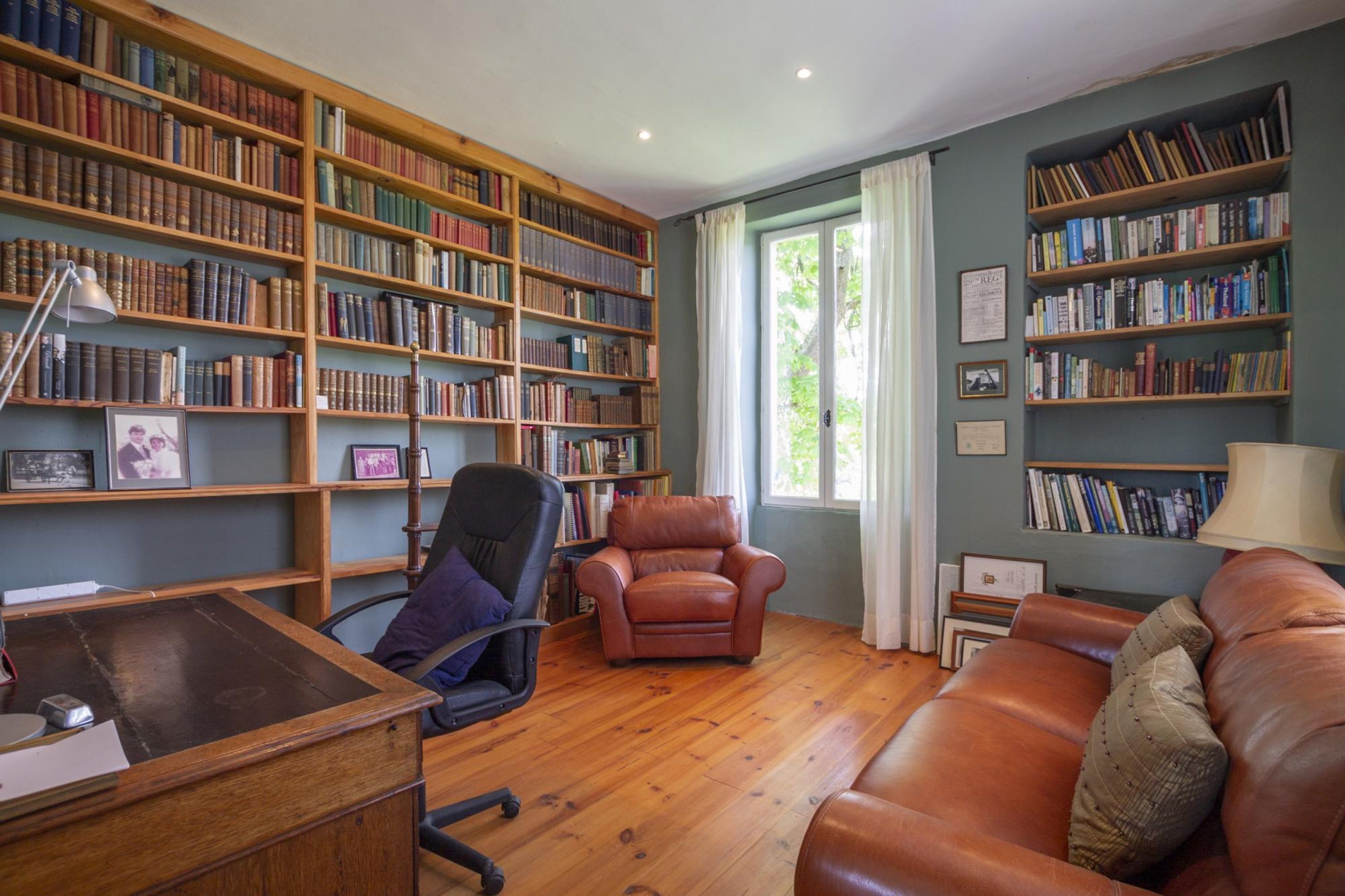 A library with wooden bookshelves, a leather armchair, a wooden desk, and a leather sofa.