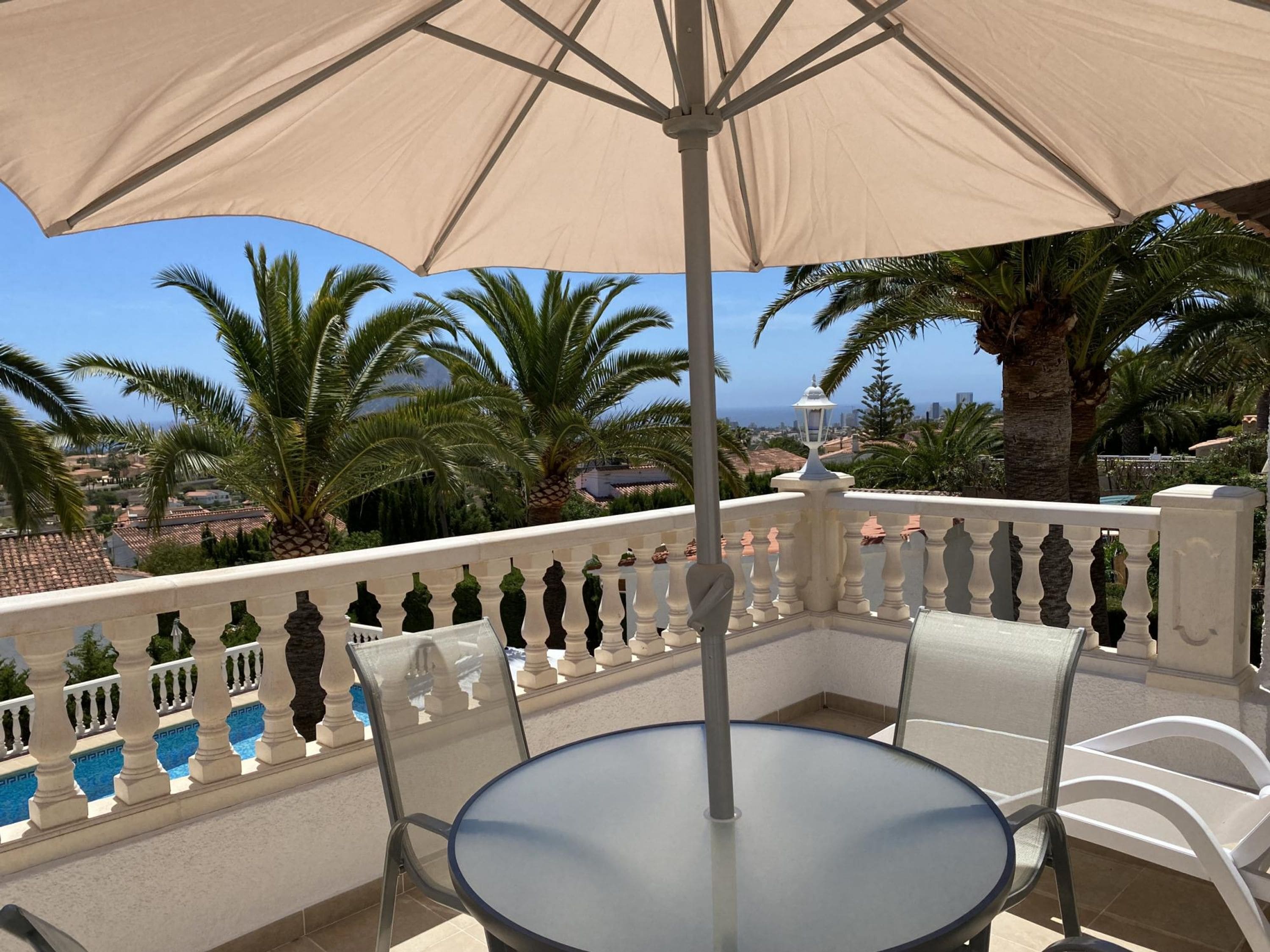 A balcony with a table and chairs, a beige umbrella, and a view of palm trees and a cityscape.