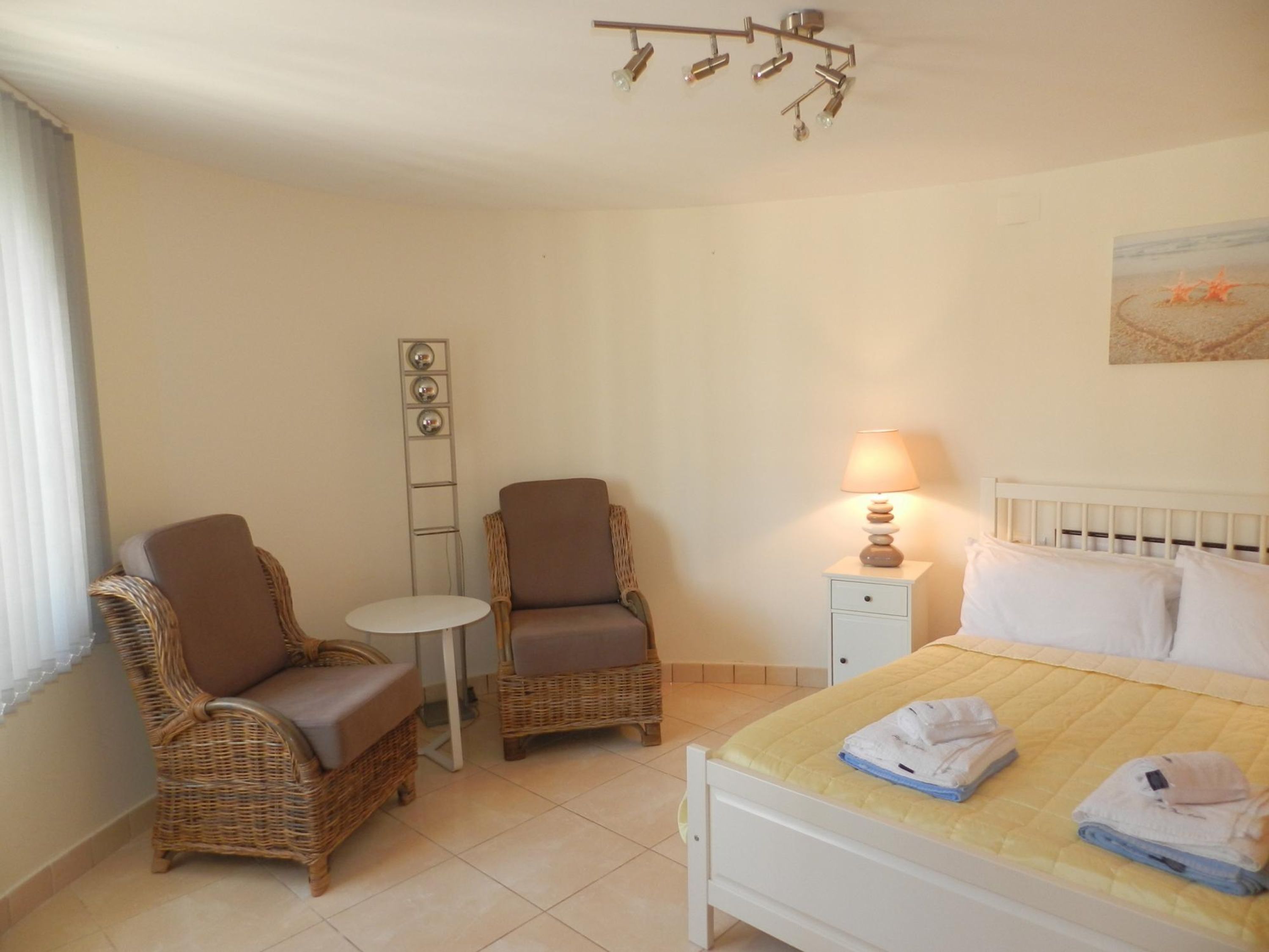 A bedroom with a bed, two wicker chairs, a small table, a bedside lamp, and a wall-mounted light fixture.
