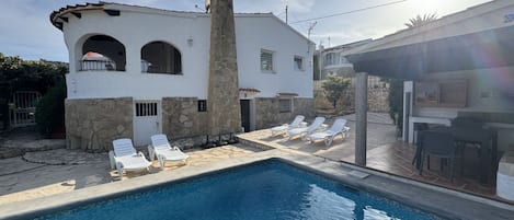 A swimming pool with white lounge chairs, a stone chimney, and a white building with arched windows.