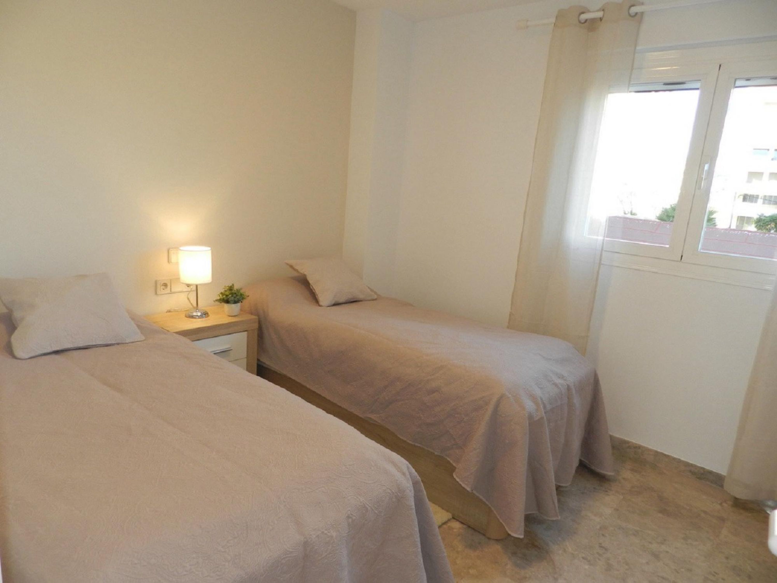 A simple, clean room with two beds, a bedside table with a lamp, and a window with curtains.