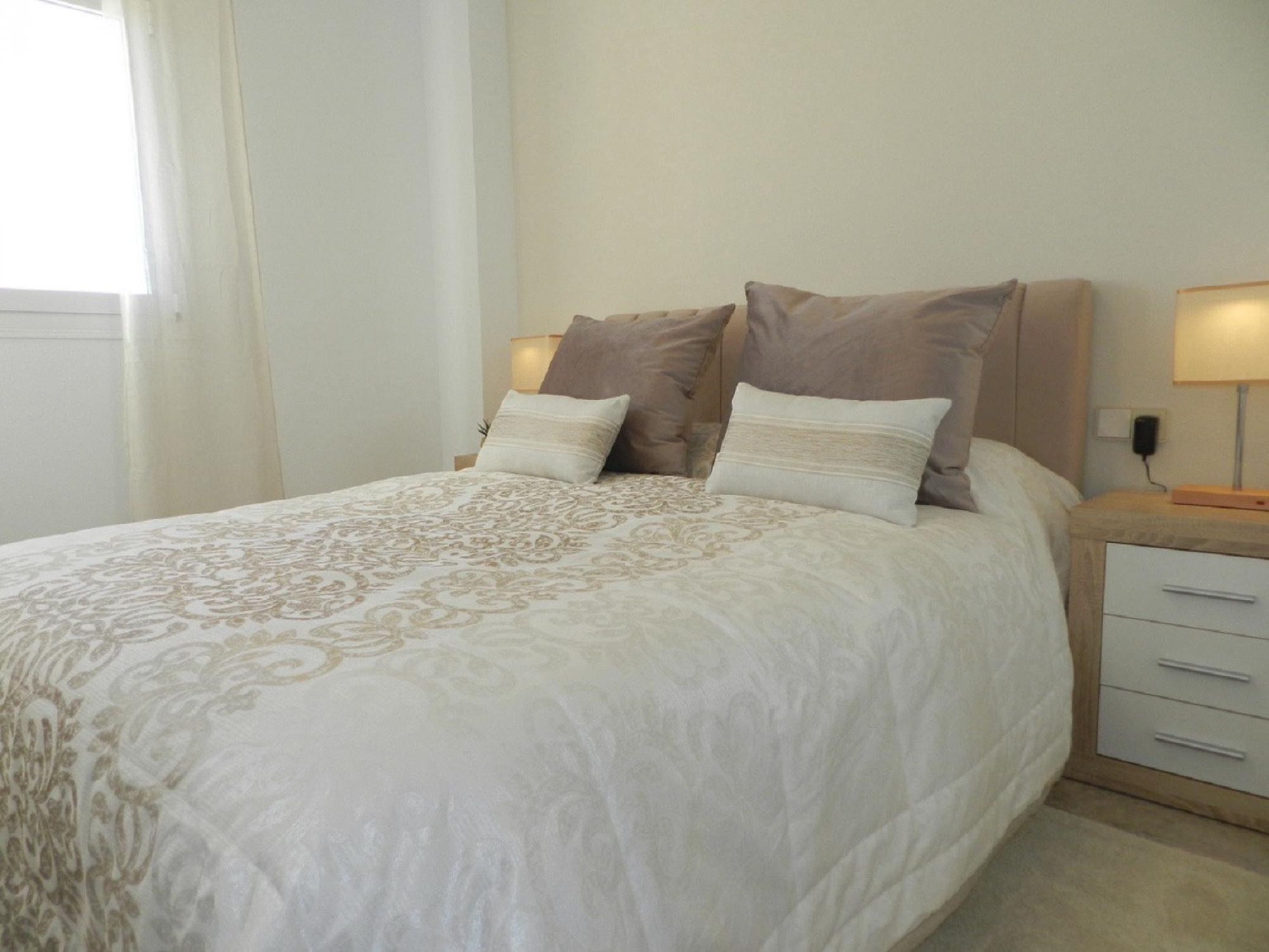 A neatly made bed with a patterned bedspread, two bedside tables with lamps, and a nightstand with a drawer.