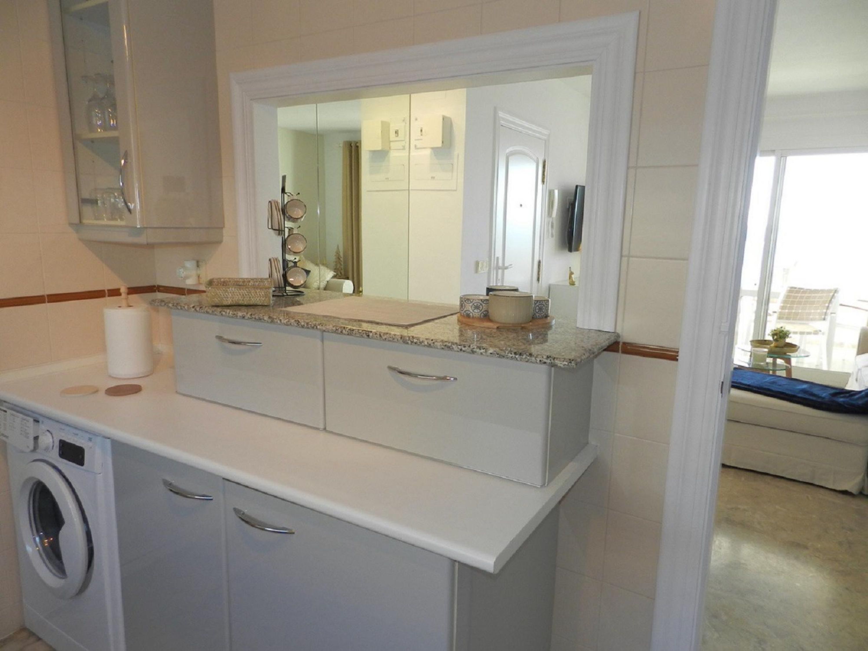 Cocina compacta con lavadora integrada, encimera con espejo y vistas al área de estar contigua.