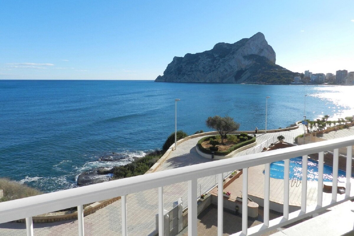 A balcony with a view of the sea, a rocky outcrop, and a cityscape in the distance.