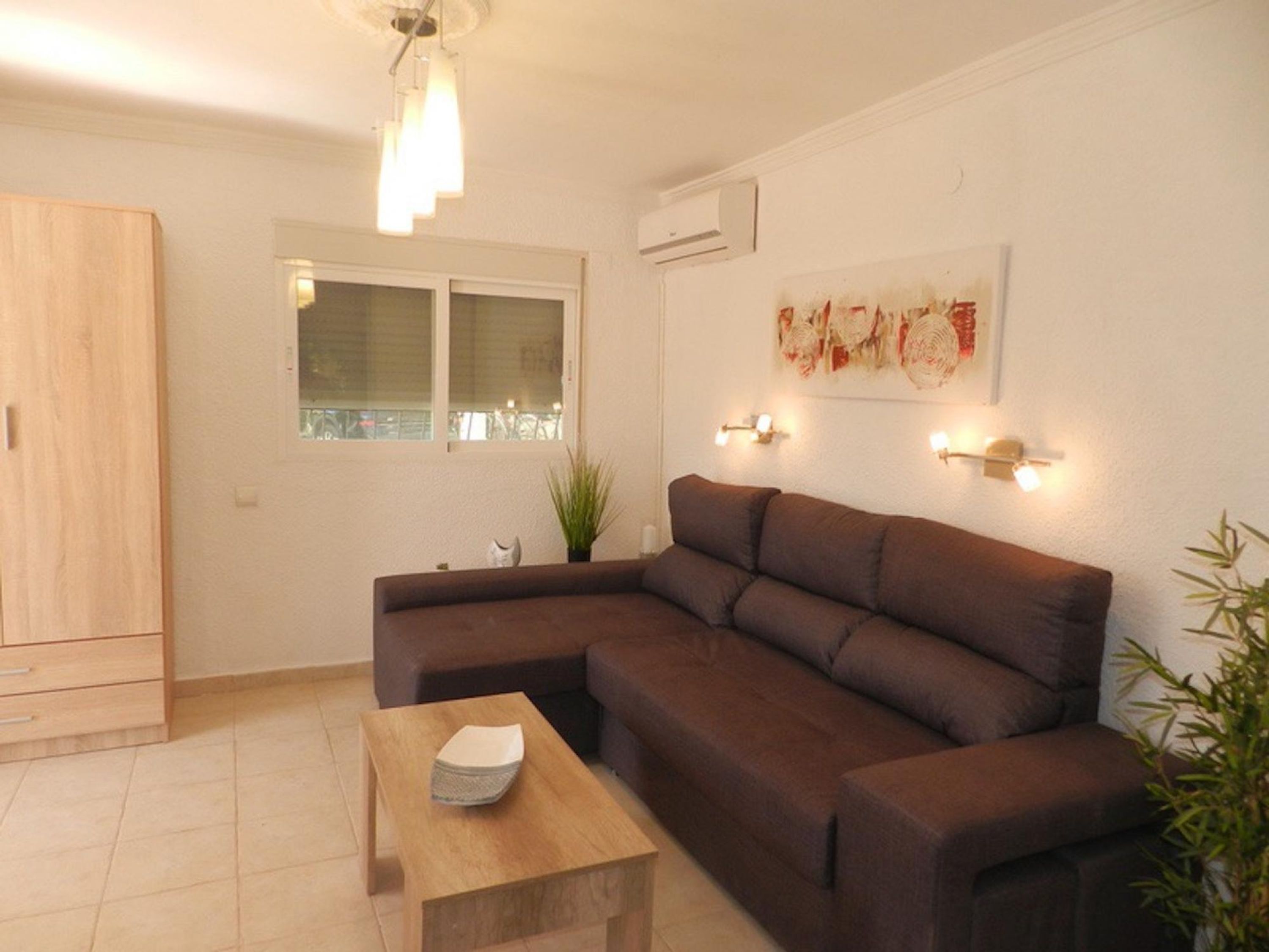 A living room with a brown sectional sofa, a wooden coffee table, a window with blinds, an air conditioner, and wall-mounted lighting.