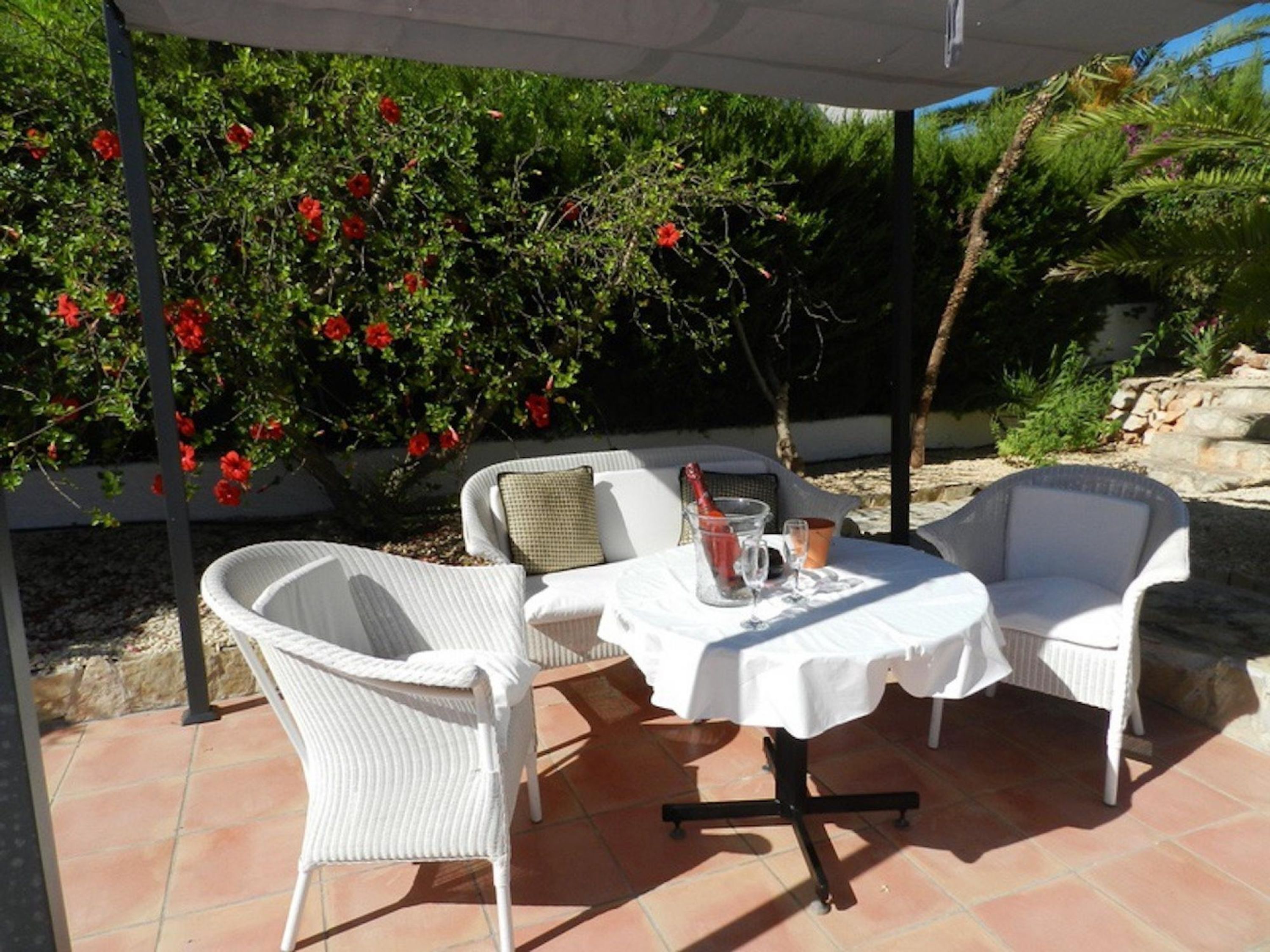 Een terras buiten met witte rieten meubels, een tafel met een witte tafelkleed en rode bloemen op de achtergrond.