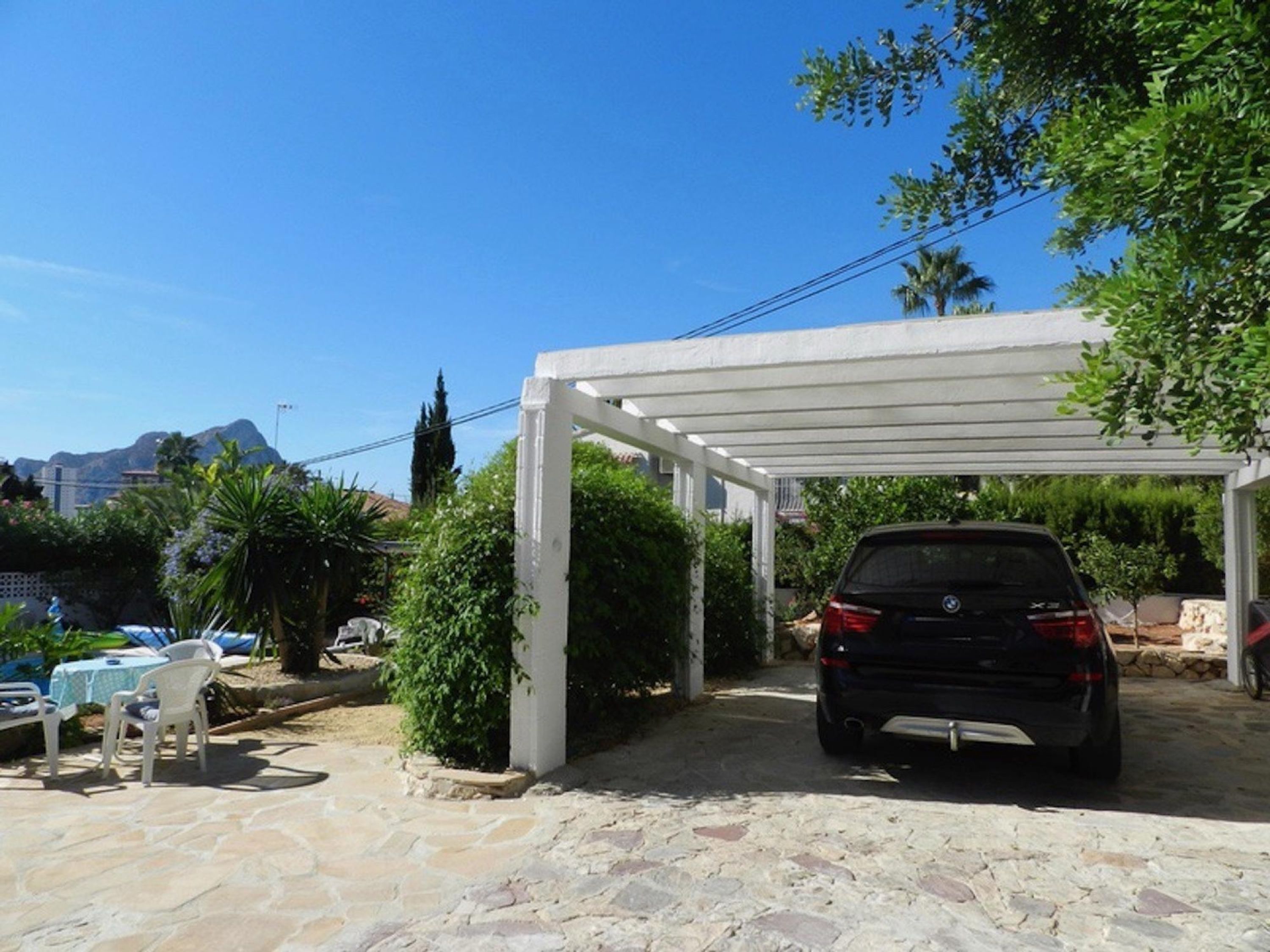 A covered outdoor area with a car parked under a white pergola, stone flooring, and greenery.