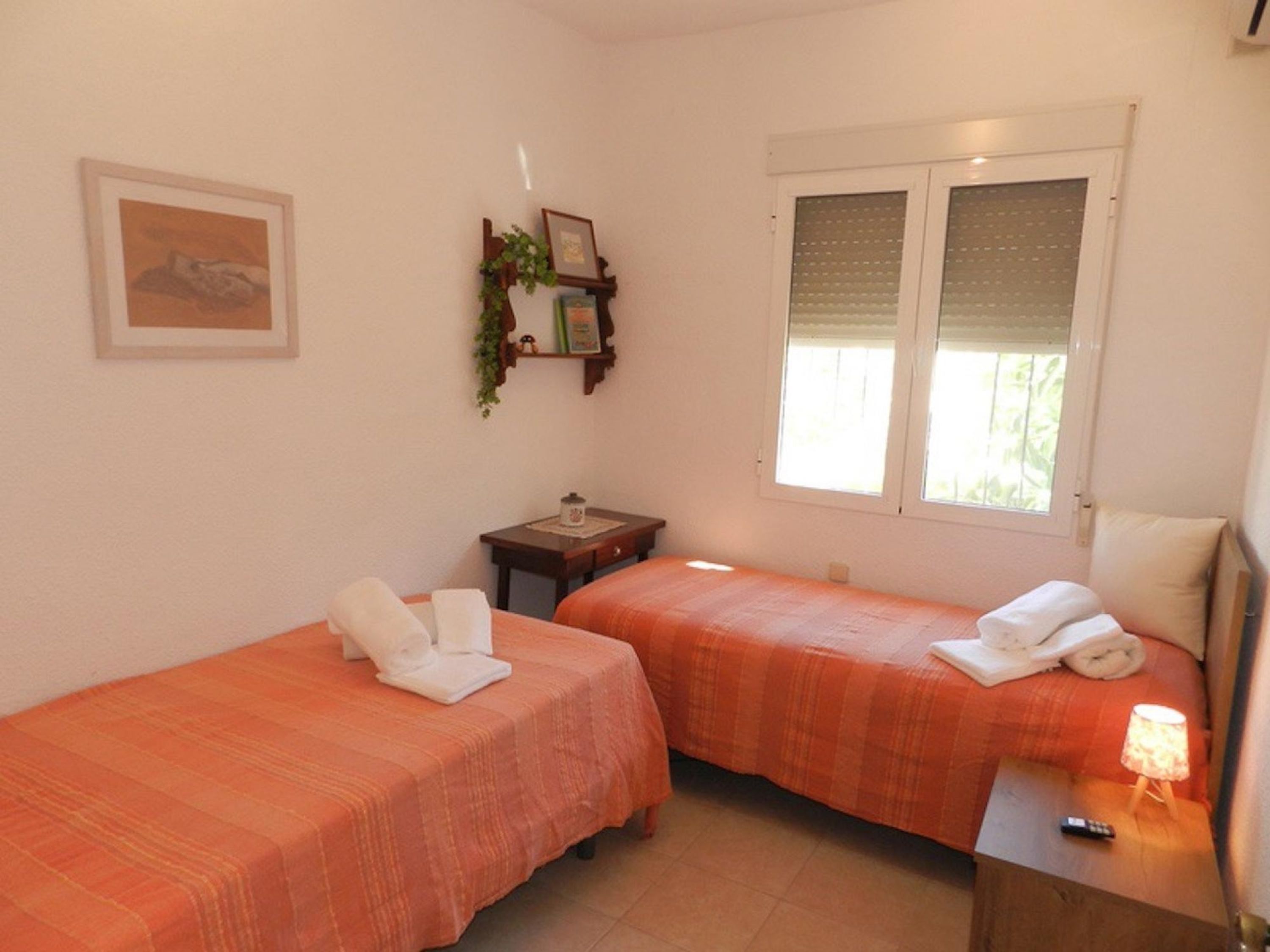 A room with two beds, each with a white towel and an orange bedspread. There is a small table with a lamp, a window with blinds, and a framed picture on the wall.