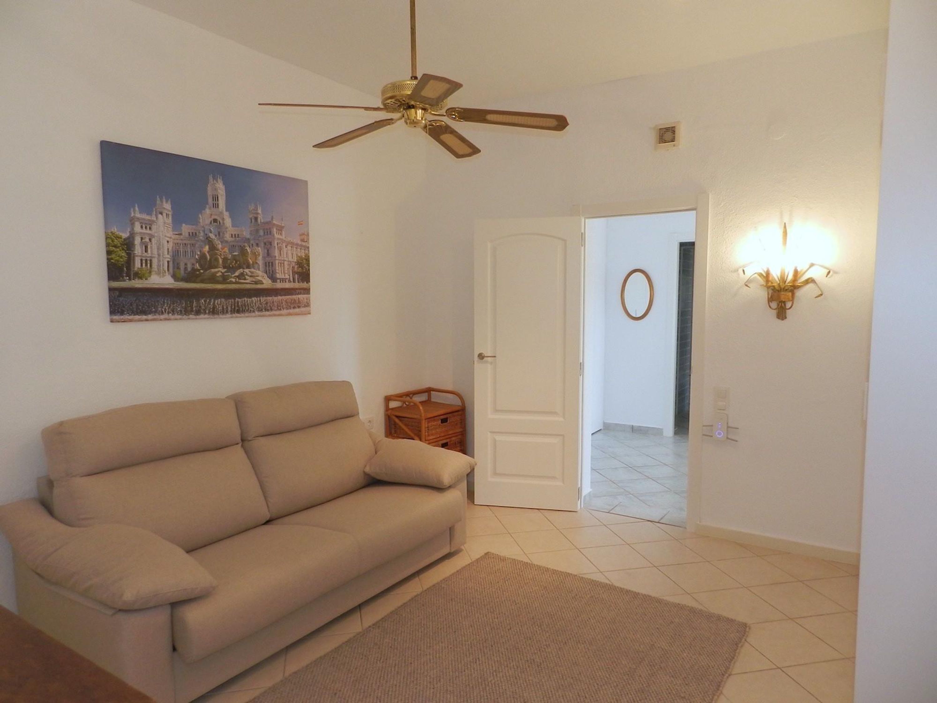 A living room with a beige sofa, a ceiling fan, a framed picture on the wall, a door leading to another room, and a wall-mounted light fixture.