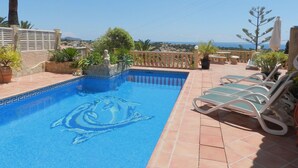 Una piscina con diseño de delfín, rodeada de sillas de descanso y plantas en macetas, con vistas al mar.