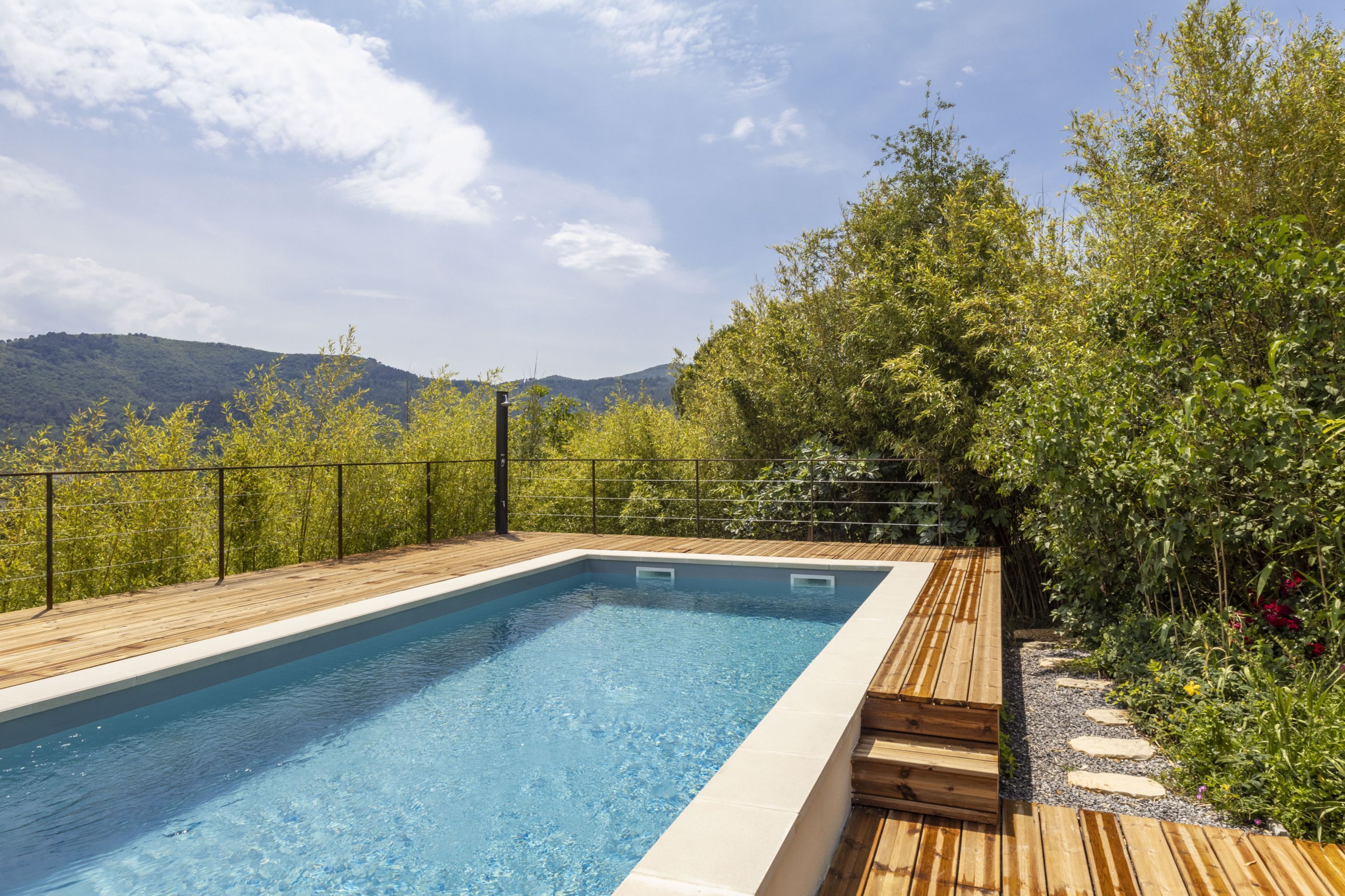 Una piscina rectangular con cubierta de madera y valla metálica, rodeada de exuberante vegetación y montañas al fondo.