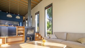 A modern living room with a wooden coffee table, a beige sofa, and a TV on a wooden stand. There are large windows providing natural light and a view of greenery outside. - La Maison des Chataigniers (Meyrannes)