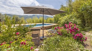 A poolside area with a wooden deck, a lounge chair, and a large umbrella. - La Maison des Chataigniers (Meyrannes)