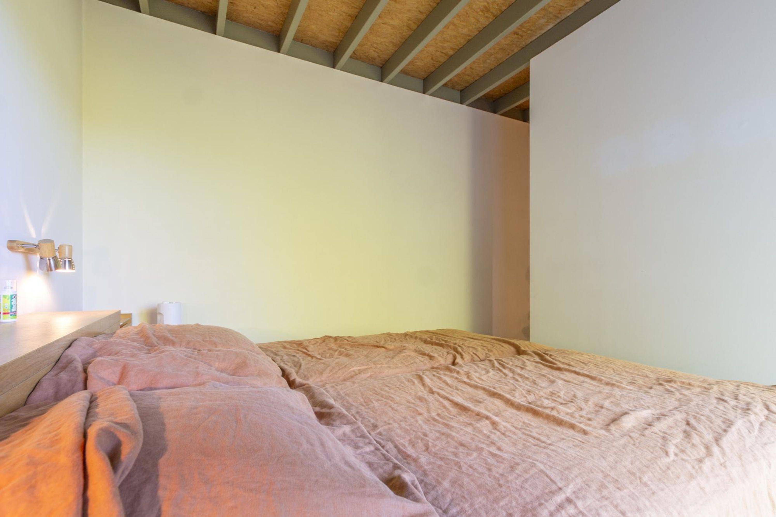 Une chambre avec une tête de lit en bois, un lit recouvert d’une couverture rose, une table de chevet avec une lampe et un mur blanc.