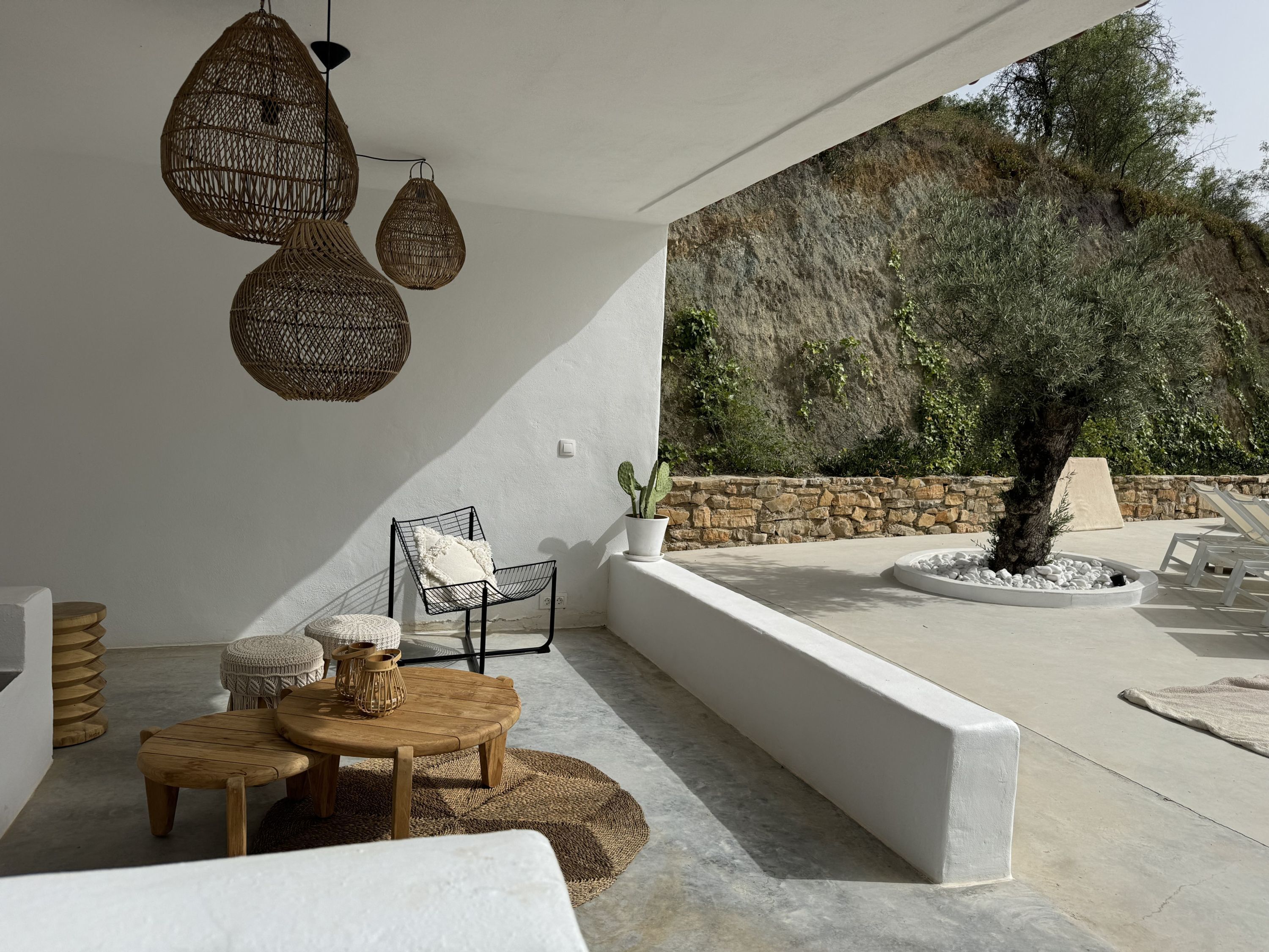 A sunlit outdoor seating area with wicker pendant lights, a stone wall, and a view of a rocky hillside.