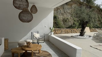 Zona d'estar a l'aire lliure amb llum solar, llums penjants de vímet, una paret de pedra i vista a un turó rocos.