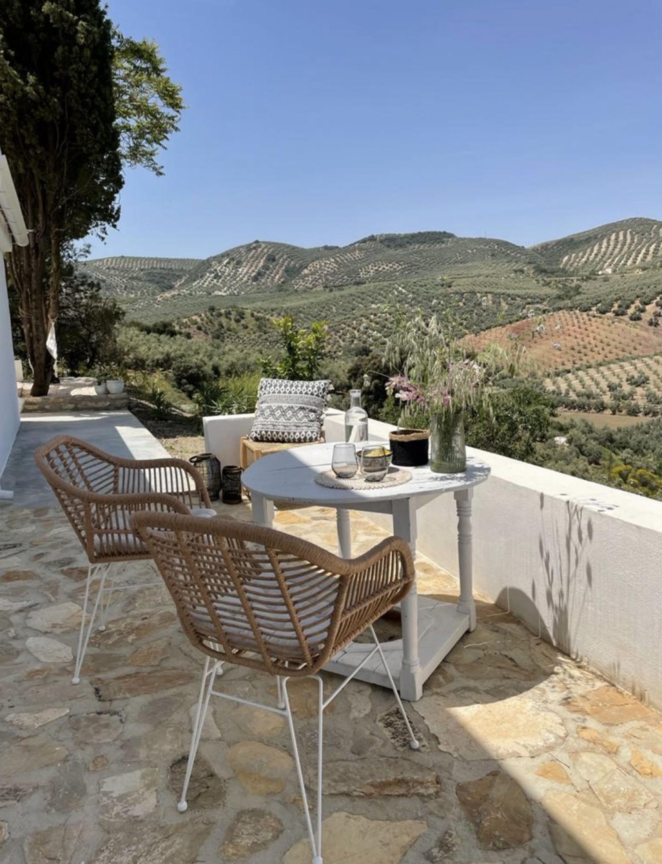 A terrace with wicker chairs and a table, overlooking a mountainous landscape.