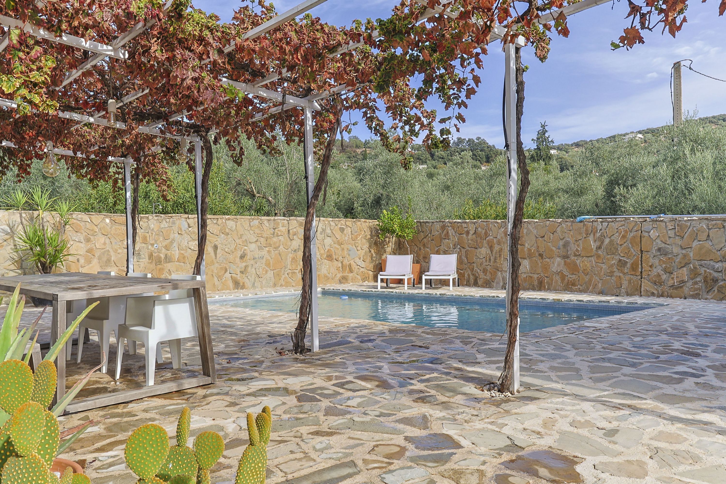 Zona de piscina con suelo de piedra, una pérgola, una mesa con sillas y vistas a una colina.