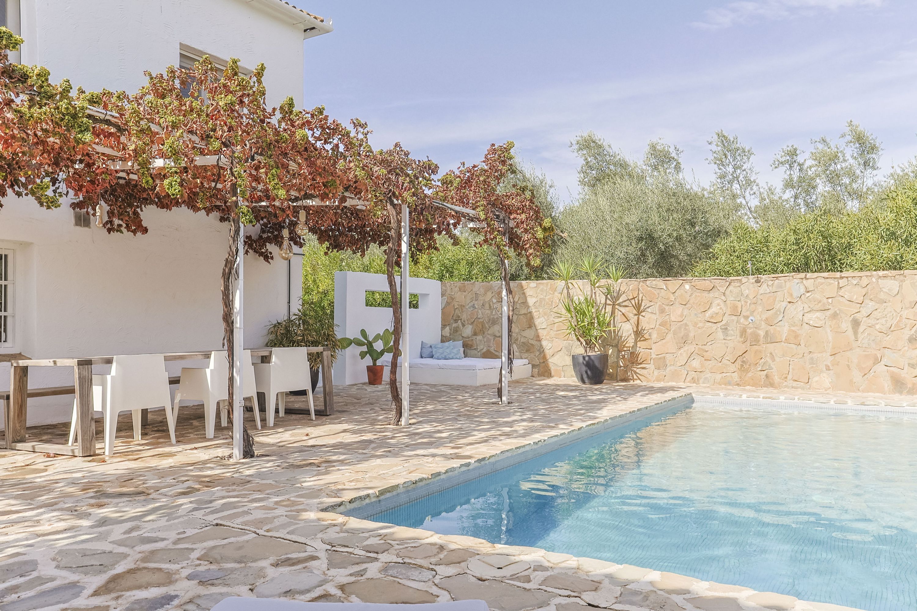 Zona de piscina con suelo de piedra, mobiliario blanco y una pérgola cubierta de hiedra de hojas rojas.