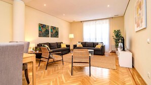 A modern living room with a herringbone floor, two sofas, a dining table, and a large window with sheer curtains.