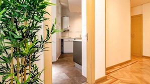 A narrow hallway leading to a kitchen with white appliances and a bamboo plant.