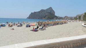 A beach with sun loungers and umbrellas, a rocky outcrop in the background, and buildings along the shore. - Apartamento Luise Para 4 Personas (Calpe)