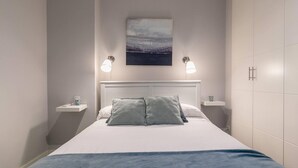 Ein ordentlich gemachtes Bett mit blauer Decke, zwei Nachttische mit Lampen, ein Wandbild und ein weißer Kleiderschrank.