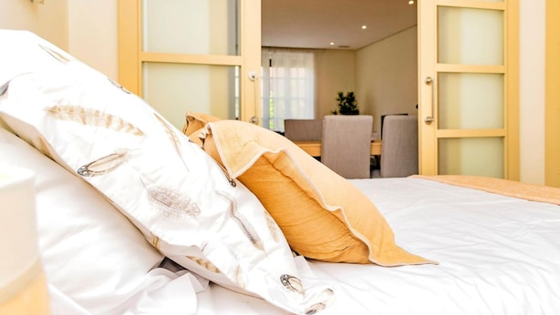 A neatly made bed with white and yellow bedding, a visible door leading to a dining area, and a small table with chairs. - Ddl20 I 2A in Madrid (Madrid)