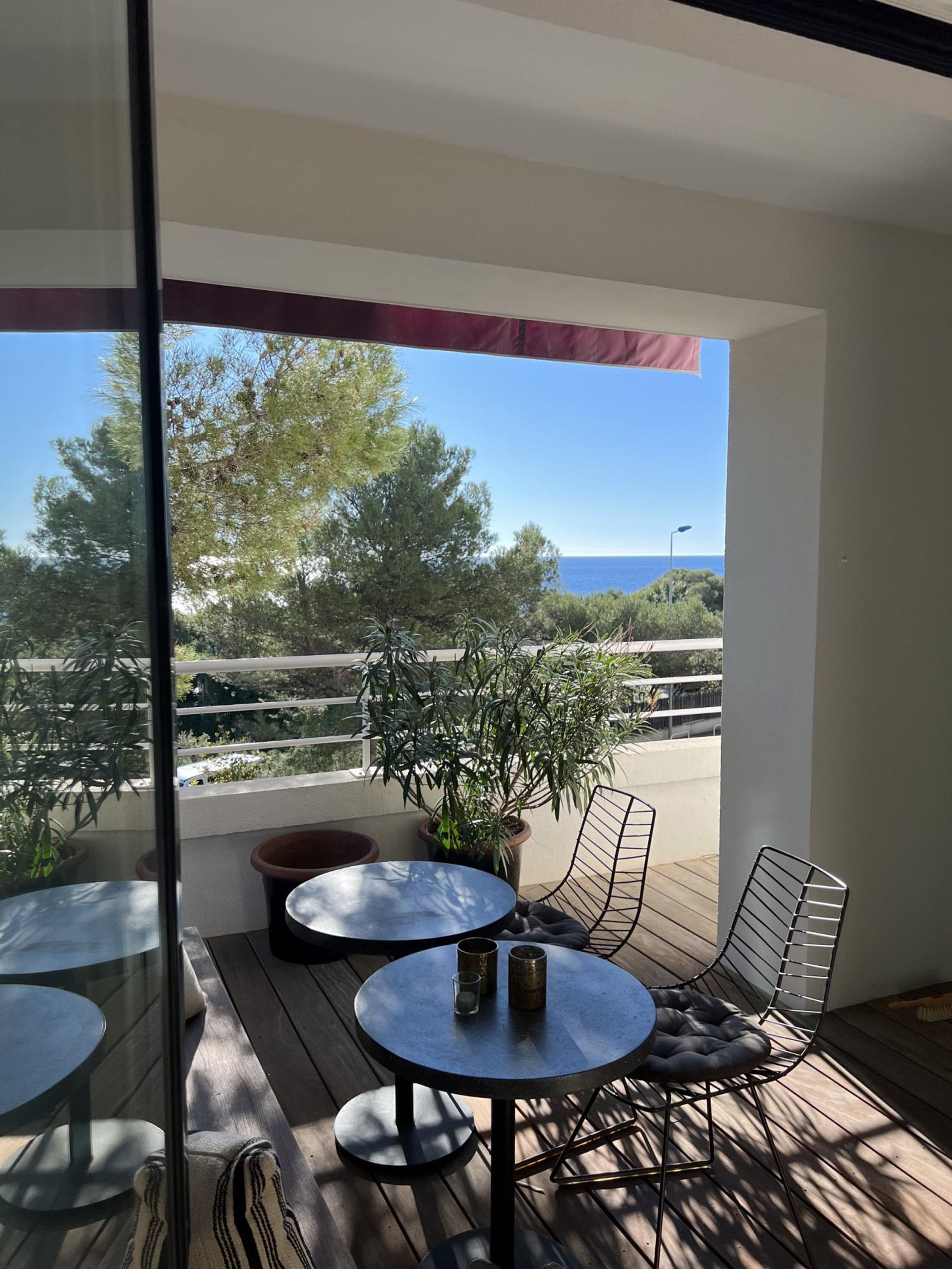 A balcony with a table and chairs, a potted plant, and a view of the sea and trees.