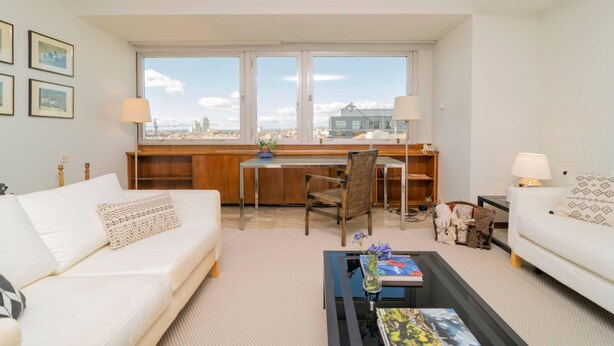 Un salon moderne comprenant un canapé, une table basse, un bureau et une chaise. Il y a deux fenêtres donnant sur la ville.