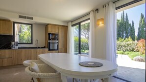 A modern kitchen with wooden cabinets, a white dining table, wicker chairs, and a view of a garden through large windows. - Villa Tica in Malauc ne (Malaucene)