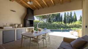 An outdoor kitchen with a grill, dining table, and chairs, overlooking a swimming pool and lush greenery. - Villa Tica in Malauc ne (Malaucene)