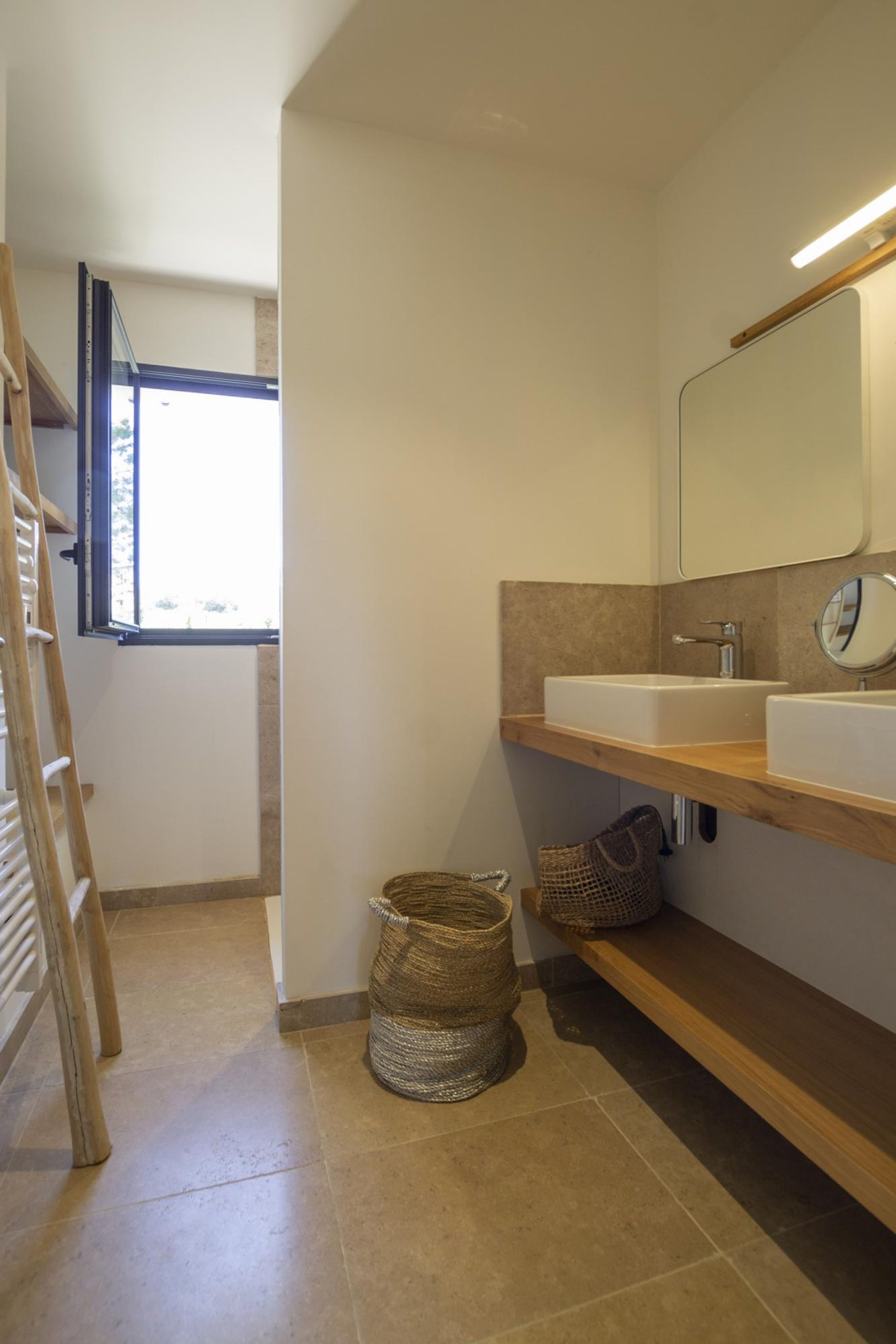 A modern bathroom with two sinks, a wooden shelf, a large mirror, and a window.