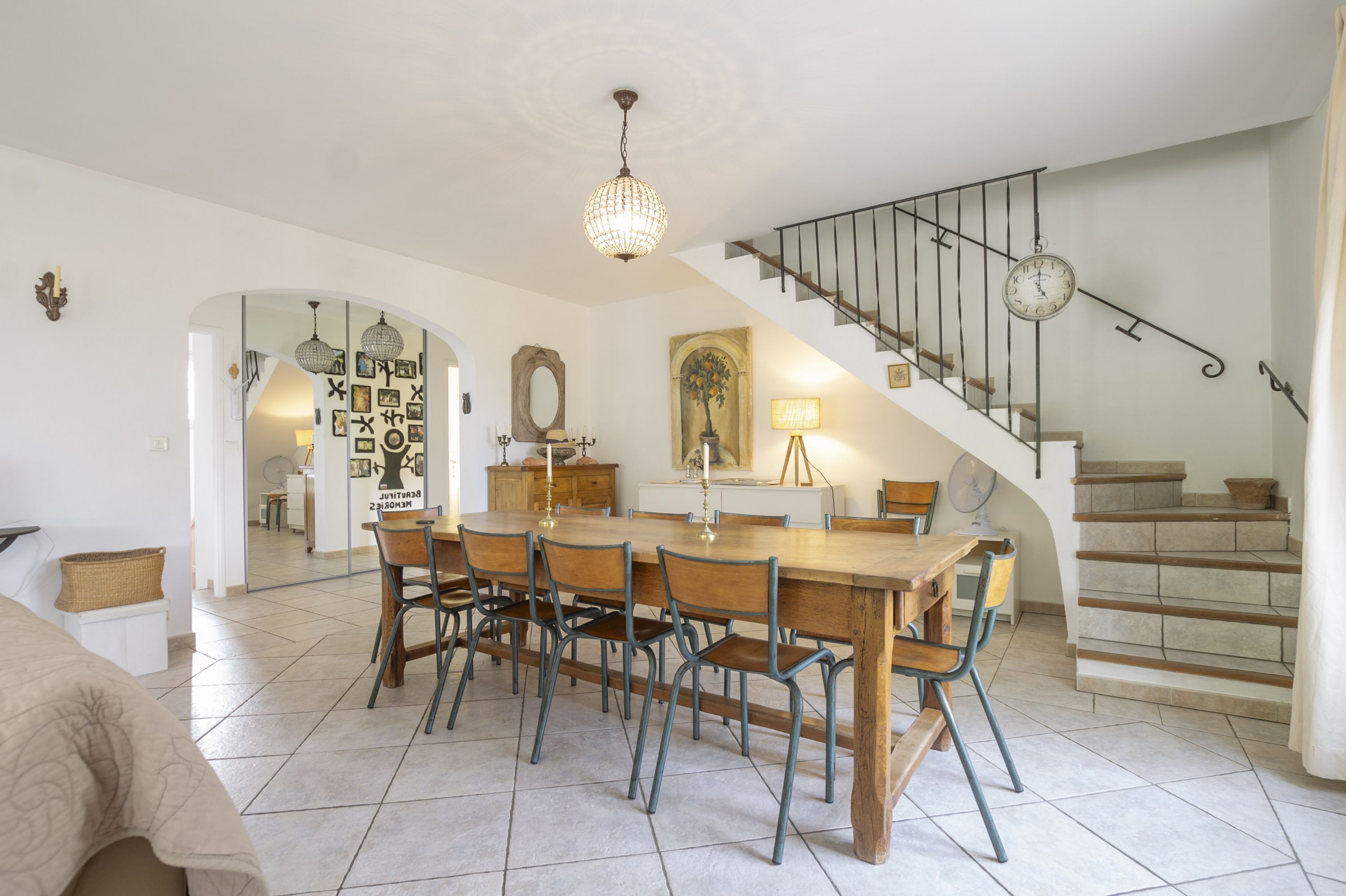 A spacious dining area with a large wooden table and chairs, a chandelier, and a staircase with a clock.