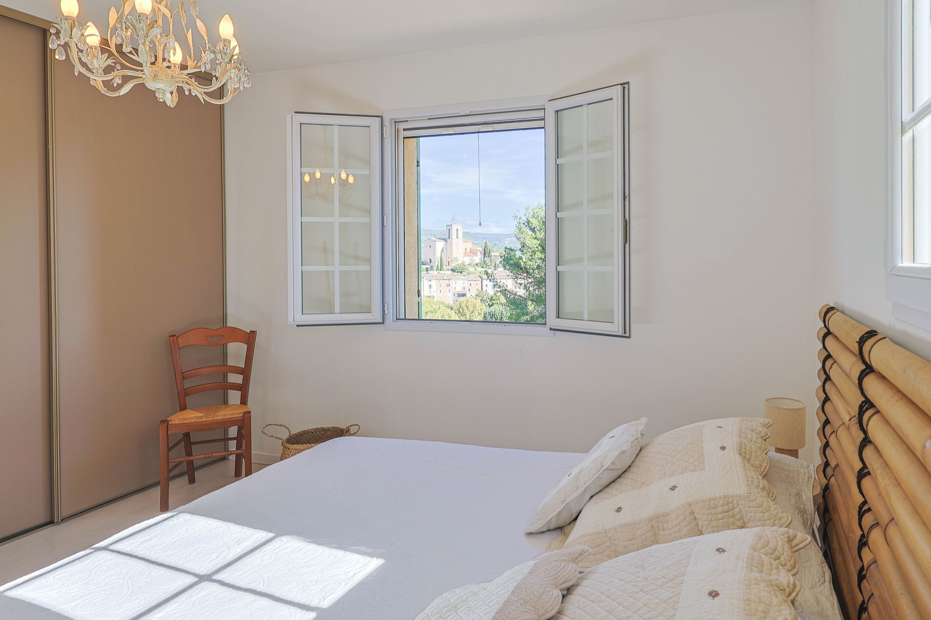 Une chambre avec un grand lit, une chaise en bois, une fenêtre dont les volets sont ouverts et un lustre.