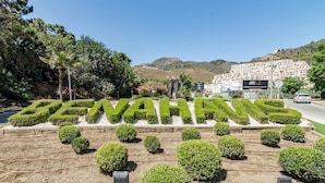 A landscaped area with the word "BENAHUIS" formed by trimmed bushes, surrounded by small trees and shrubs, with a road and buildings in the background. - RI B2a- Relaxed Modern Flat in Benahavis (Benahavis)