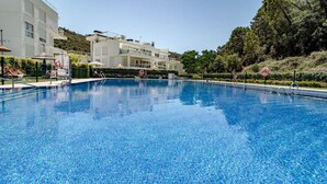 A clear blue pool with a tiled bottom, surrounded by a white building with balconies and a hilly backdrop. - RI B2a- Relaxed Modern Flat in Benahavis (Benahavis)