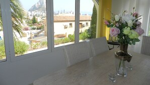 A dining area with a table and chairs, a vase of flowers, and a view of a cityscape through large windows. - Villa Fig Tree Para 4 Personas (Calpe)