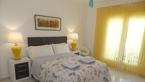 A neatly made bed with a floral bedspread, two bedside lamps, a nightstand with drawers, a chair, and a window with yellow curtains. - Villa Fig Tree Para 4 Personas (Calpe)