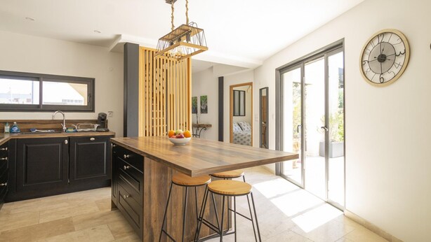 A modern kitchen with a wooden island, bar stools, a hanging light fixture, and a large window. - Maison des Fleurs in Ved ne (Vedene)