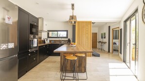 A modern kitchen with dark cabinetry, a central island with stools, a hanging light fixture, and a view of the outdoors. - Maison des Fleurs in Ved ne (Vedene)