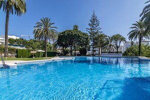 Une piscine d’un bleu éclatant, entourée de palmiers et de verdure, avec des bâtiments en arrière-plan.