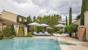 A pool area with lounge chairs and umbrellas, surrounded by greenery and a building. - Villa Ambre - Private Pool and Magnificent View (Digne-les-Bains)