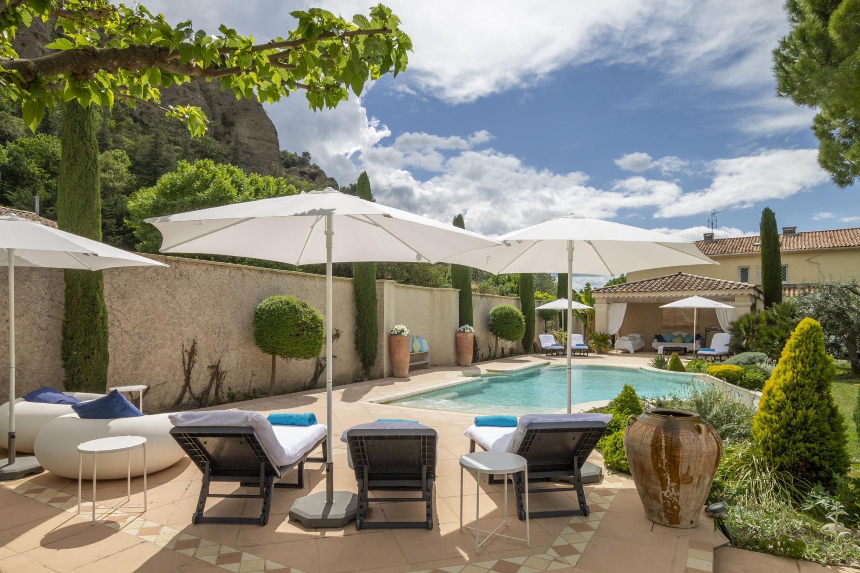 A poolside area with lounge chairs and umbrellas, surrounded by greenery and a mountain backdrop.