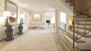 Un'ampia sala con pavimento rivestito di moquette, un tavolino consolle con piano in vetro, lampade e un grande specchio, e una scala con ringhiera in ferro battuto.